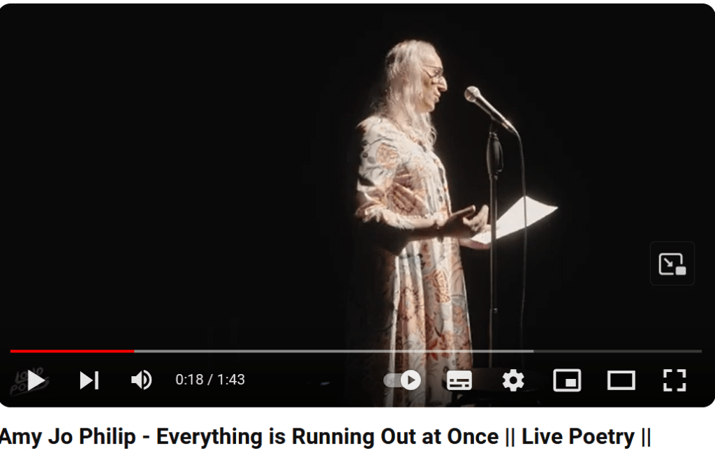 Still from a video of Amy Jo, a trans woman, reading a poem. Amy Jo is shown in profile wearing a white dress with a large floral pattern and has a sheaf of papers in one hand. Click the image to go to the video. 