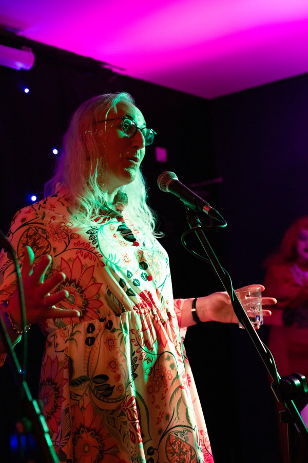 Amy Jo, a trans woman in a floral dress, standing a microphone with her arms wide and her hands spread.